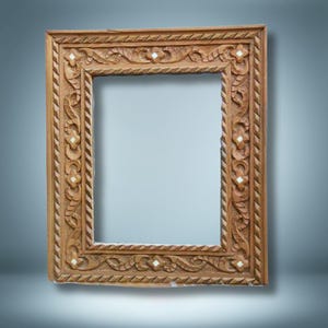 Puede incluir: Un marco de fotos rectangular de madera marrón claro con intrincados patrones florales y geométricos tallados. El marco tiene un borde detallado y pequeños acentos incrustados blancos, creando un diseño ornamental.