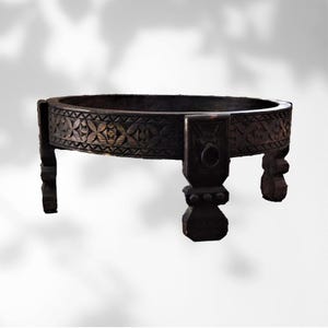 May include: A dark brown, low, round wooden table with carved floral patterns around the rim. The table has four short, sturdy legs and a decorative ring pull on one side. The table is approximately 12 inches high.