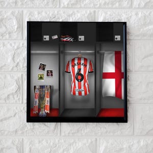 May include: A framed display featuring a soccer jersey with the name "BASHAM" and the number "6" on the back, hanging in a locker. The display includes a trophy, photos, and a pair of cleats. An English flag is also visible.