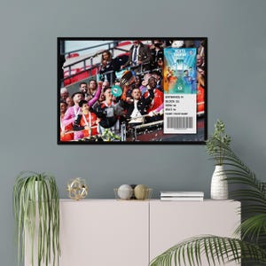 May include: A framed print depicting a sports celebration with a trophy and confetti. A ticket with the text "VERTU TROPHY" and other details is visible. The print is displayed on a wall above a pink cabinet with decorative items.