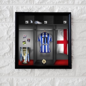 Könnte beinhalten: Gerahmte Präsentation von Fußball-Erinnerungsstücken. Enthält ein blau-weiß gestreiftes Trikot mit der Nummer 11 und dem Namen "WINDASS", eine Trophäe, einen Fußballschuh und eine englische Flagge. Die Kulisse ist eine Umkleidekabine.