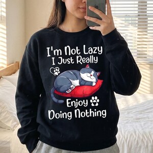 May include: A black long-sleeve sweatshirt featuring a graphic of a grey and white cat sleeping on a red pillow. The text reads, "I'm Not Lazy I Just Really Enjoy Doing Nothing," accompanied by two small white paw print icons.