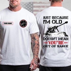 May include: A white t-shirt featuring a front design with a sniper scope graphic and a box for a name, and a back design showing a sniper with the text JUST BECAUSE I'M OLD DOESN'T MEAN YOU'RE OUT OF RANGE. Both sleeves feature a small American flag.