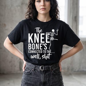 May include: A black short-sleeved t-shirt featuring a white graphic of a skeleton and the text "The KNEE BONE'S CONNECTED TO THE.... well, shit". The skeleton is depicted in a dancing pose with one leg raised, positioned next to the bold white lettering.