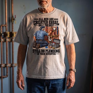 May include: A light gray t-shirt featuring a graphic of a plumber kneeling before a brick wall with pipes, a toolbox, and the text: ALL MEN ARE CREATED EQUAL ONLY THE BEST STILL DO PLUMBING IN THEIR SEVENTIES. The design includes a pressure gauge.
