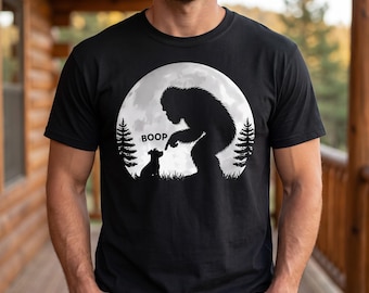 May include: A black short-sleeved t-shirt featuring a white graphic of a large creature and a small dog standing in a forest under a full moon. The creature is reaching out to touch the dog's nose, with the word BOOP printed above the dog.