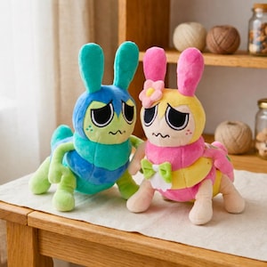 Puede incluir: Dos peluches h&iacute;bridos de oruga y conejo. Uno es azul y verde, el otro rosa y amarillo. Ambos tienen ojos grandes, bocas ca&iacute;das y orejas de conejo. El rosa y amarillo tiene una flor y un lazo.