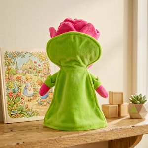 Puede incluir: Un peluche con forma de rosa rosa con una capa verde, de pie sobre una estanter&iacute;a de madera. Un libro infantil con ilustraciones y bloques de madera tambi&eacute;n est&aacute;n en la estanter&iacute;a. Tambi&eacute;n se ve una peque&ntilde;a planta suculenta en una maceta geom&eacute;trica.