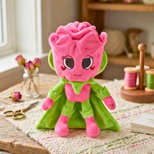 Puede incluir: Un peluche con forma de rosa rosa con una cara sonriente, una capa verde y detalles verdes a juego. El juguete est&aacute; colocado sobre una superficie texturizada de color crema. El juguete tiene grandes ojos negros y detalles en forma de coraz&oacute;n.