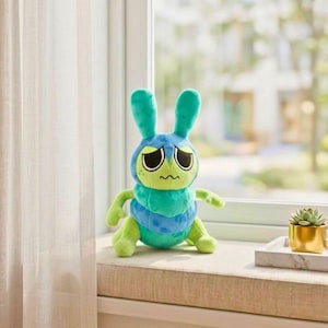 Puede incluir: Un peluche con forma de h&iacute;brido de oruga y conejo azul y verde con una expresi&oacute;n triste. El juguete tiene ojos grandes y ca&iacute;dos, orejas largas y un cuerpo segmentado. Est&aacute; colocado en un alf&eacute;izar cerca de una peque&ntilde;a suculenta en una maceta dorada.