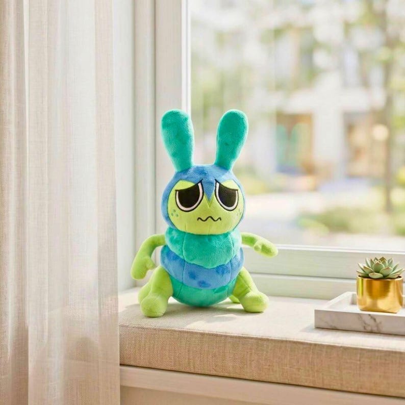 Puede incluir: Un peluche con forma de h&iacute;brido de oruga y conejo azul y verde con una expresi&oacute;n triste. El juguete tiene ojos grandes y ca&iacute;dos, orejas largas y un cuerpo segmentado. Est&aacute; colocado en un alf&eacute;izar cerca de una peque&ntilde;a suculenta en una maceta dorada.