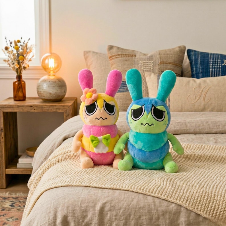 Puede incluir: Dos peluches con forma de conejos con expresiones tristes. Uno es rosa y amarillo con una flor, el otro es azul y verde. Tienen orejas largas y est&aacute;n sentados en una cama con ropa de cama de tonos neutros.
