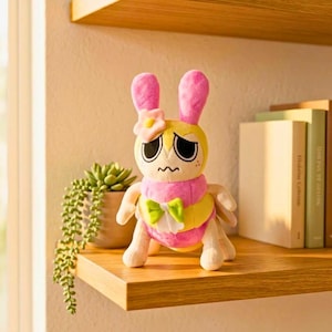 Puede incluir: Un peluche con forma de conejo con orejas rosas, cuerpo amarillo y bufanda rosa. Tiene una flor en la cabeza y un lazo verde. El juguete est&aacute; sobre una estanter&iacute;a de madera.
