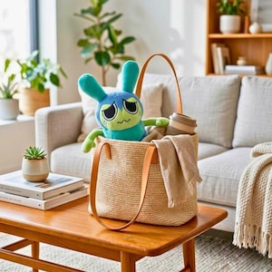 Puede incluir: Un bolso tote tejido con asas de cuero marr&oacute;n y un peluche de conejo azul y verde en su interior. Una taza de caf&eacute; reutilizable y una manta beige tambi&eacute;n est&aacute;n en la bolsa. La bolsa est&aacute; sobre una mesa de madera.