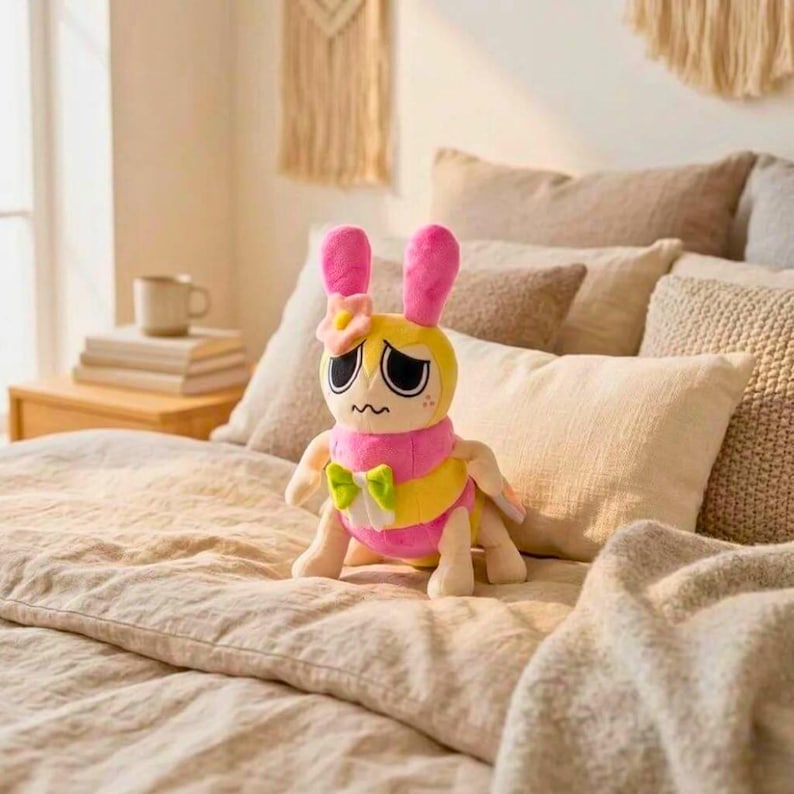 Puede incluir: Un peluche con forma de h&iacute;brido insecto-conejo amarillo y rosa con ojos grandes y una flor en la cabeza. El juguete tiene orejas rosas, una bufanda rosa y un lazo verde. Est&aacute; colocado sobre una cama con ropa de cama beige.