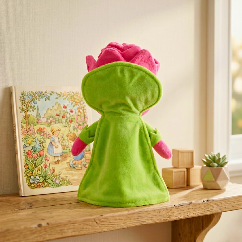 Puede incluir: Un peluche con forma de rosa rosa con una capa verde, de pie sobre una estanter&iacute;a de madera. Un libro infantil con ilustraciones y bloques de madera tambi&eacute;n est&aacute;n en la estanter&iacute;a. Tambi&eacute;n se ve una peque&ntilde;a planta suculenta en una maceta geom&eacute;trica.
