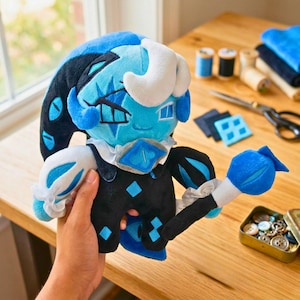May include: A plush doll with a blue and black jester-like design, holding a blue paintbrush. The doll has white accents and a diamond-shaped collar. The doll is made of soft fabric and has a whimsical appearance.