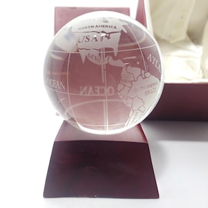Puede incluir: Un globo terráqueo de cristal transparente grabado con continentes y nombres de países, incluyendo "USA" y "Mexico". El globo se encuentra sobre una base trapezoidal de color burdeos. El globo está dentro de una caja burdeos.