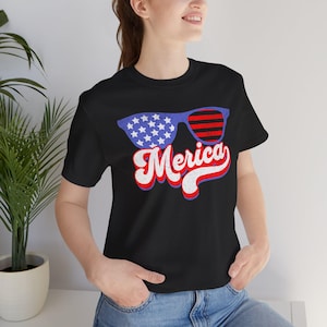 May include: Black t-shirt with a graphic of sunglasses with a blue frame and red and white stripes and stars. The word "Merica" is written in red, white, and blue script below the sunglasses.