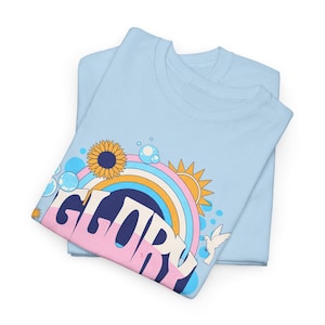 May include: Two pale blue t-shirts are stacked. The top one has a colourful graphic design featuring a rainbow, sunflower, sun, bubbles, and the word "GLORY" in a retro font. A white dove is also included in the design.
