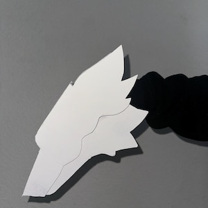 May include: A white paper cutout shaped like a stylized animal head with pointed ears and a snout, featuring a distinct layered design. The cutout is held by a black fabric sleeve, showcasing a clean, sharp silhouette against a neutral background.