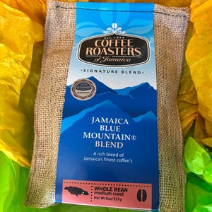 Op de afbeelding: Een jute koffiezak met een blauw etiket met de tekst "Jamaica Blue Mountain® Blend" en "Coffee Roasters of Jamaica". Het etiket bevat ook een berg-afbeelding en geeft aan dat de koffie hele boon, medium roast en 227g is.