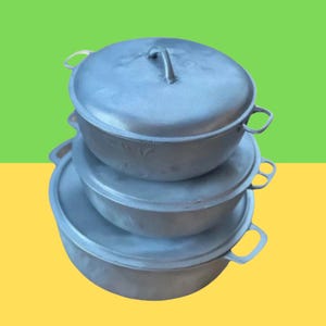 Puede incluir: Un conjunto de tres ollas de aluminio apiladas, con tapas. Las ollas son de diferentes tamaños, con cuerpos redondeados y asas de bucle. Las tapas tienen un solo asa en la parte superior. Fondo verde y amarillo.