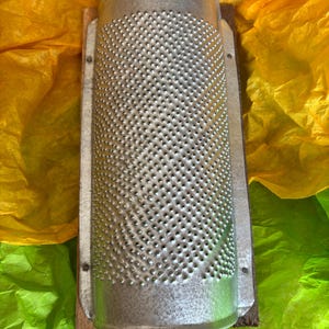 May include: A vintage metal grater with a cylindrical design and numerous small holes. The grater is mounted on a wooden base and is set against a backdrop of yellow and green tissue paper. The grater is a kitchen tool.