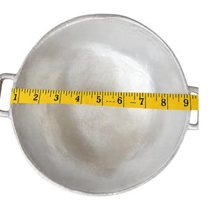 May include: A round, silver-colored metal cooking pot with two side handles. A yellow measuring tape is placed across the center of the pot, showing a diameter of approximately 9 inches from the inner edge to the opposite inner edge.