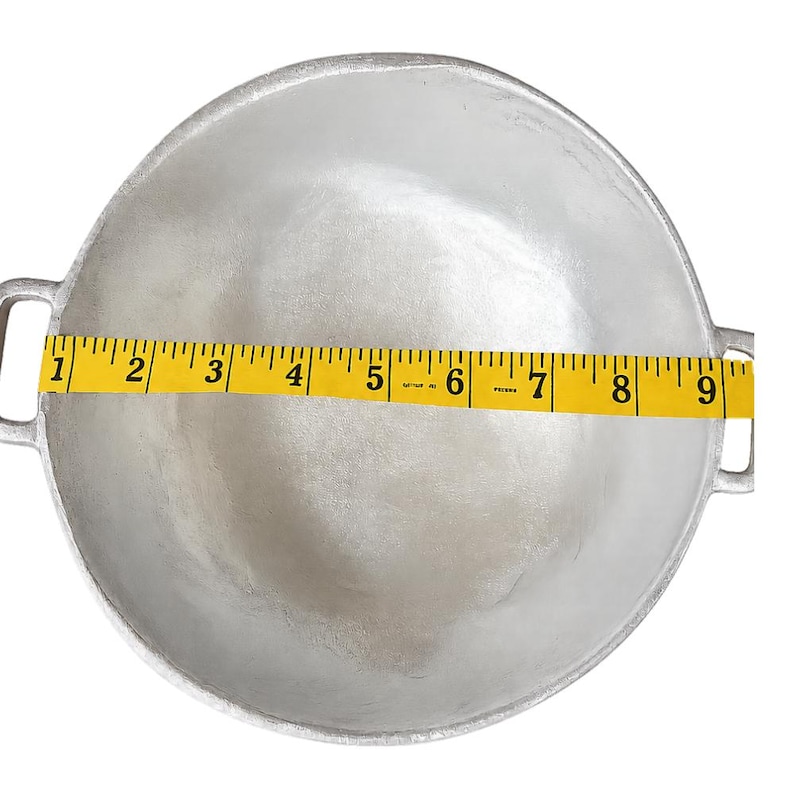 May include: A round, silver-colored metal cooking pot with two side handles. A yellow measuring tape is placed across the center of the pot, showing a diameter of approximately 9 inches from the inner edge to the opposite inner edge.