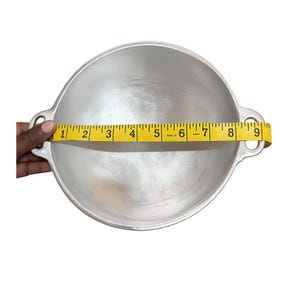 May include: A round, silver-colored metal cooking pot with two side handles. A yellow measuring tape is stretched across the center of the pot, indicating a diameter of approximately 9 inches from the outer edge of one handle to the other.