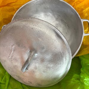 Peut inclure: Une marmite en fonte de couleur argentée avec un couvercle assorti. La marmite a deux poignées et un couvercle avec une poignée. La marmite et le couvercle sont sur une surface jaune et verte.