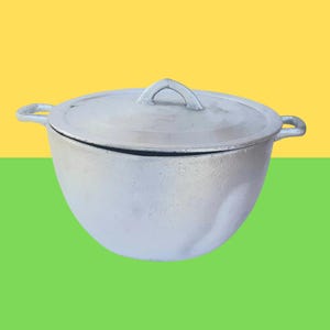 May include: A silver-colored cooking pot with a matching lid. The pot has two side handles and a loop handle on the lid. The background is divided into yellow and green sections.