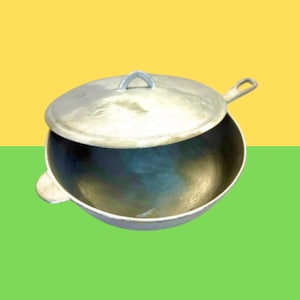 May include: A round, silver-colored metal skillet with a matching lid. The pan features a short, flat handle on one side and a small, rounded tab handle on the opposite side. The lid has a central, arched handle for lifting and a small side extension.