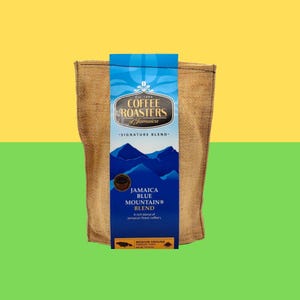 Pode incluir: Um saco de juta de café Jamaica Blue Mountain Blend. A etiqueta azul apresenta o texto "COFFEE ROASTERS of Jamaica" e "JAMAICA BLUE MOUNTAIN BLEND". A etiqueta também inclui um gráfico de montanha e as palavras "SIGNATURE BLEND".