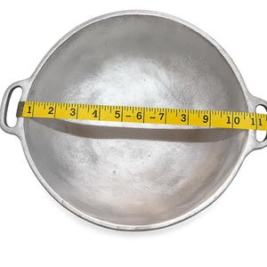 May include: A round, silver-colored metal cooking pot with two side handles. A yellow measuring tape is placed across the center of the pot, showing a diameter measurement of approximately 11 inches from one inner edge to the other.