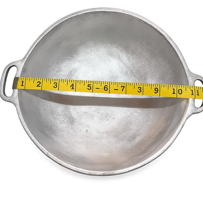 May include: A round, silver-colored metal cooking pot with two side handles. A yellow measuring tape is placed across the center of the pot, showing a diameter measurement of approximately 11 inches from one inner edge to the other.