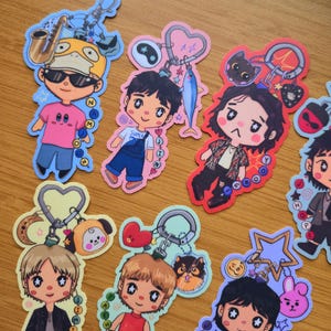 May include: A collection of die-cut stickers featuring chibi-style characters with star-shaped eyes, each attached to a colorful keychain ring with various charms including animals, musical instruments, and shapes like hearts, stars, and moons.