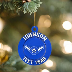May include: A blue Christmas ornament with the name "JOHNSON" in white lettering around the top edge. The center features a white emblem with a star. The bottom edge reads "TEXT, YEAR". The ornament hangs from a gold string.