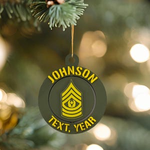 Puede incluir: Adorno navideño redondo, verde oliva, con texto amarillo que dice "JOHNSON" en la parte superior y "TEXT, YEAR" en la inferior. El centro presenta una insignia de rango militar amarilla. Una cuerda dorada está unida para colgarlo. Diámetro aproximado de 8 cm.