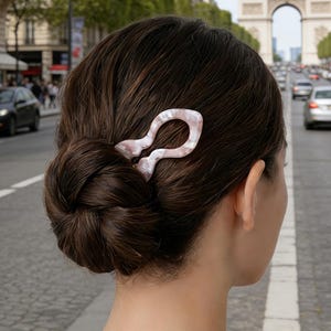 May include: A light pink and white marbled hair clip is placed in a low bun hairstyle. The hair clip has a unique, abstract shape. The background shows a city street with cars and a large archway.