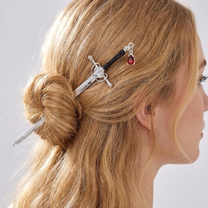 May include: Silver-toned hair stick in the shape of a sword, with a black handle and a red teardrop-shaped charm. The hair accessory is inserted into a bun of blonde hair, creating a unique and stylish look.
