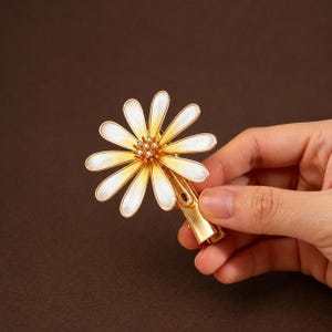 Può includere: Una molletta per capelli in metallo dorato con un ornamento a forma di margherita. Il fiore presenta dodici petali allungati con una finitura in smalto bianco che sfuma verso un centro giallo. Il cuore del fiore è decorato con piccoli cristalli.