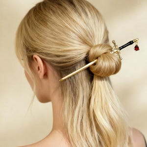 May include: A gold-colored hair stick and sword-shaped hair accessory with a black handle and a red gem. The hair accessories are placed in a blonde bun hairstyle. The image is taken from the back, showing the person's hair and neck.