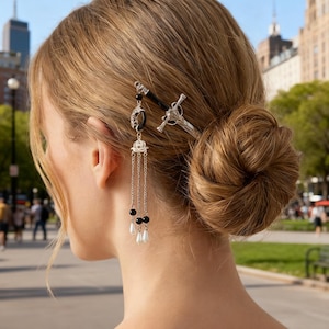 May include: A woman's hair is styled in a bun, adorned with two decorative hairpins. One is a silver sword design, and the other features black beads, dangling chains, and faux pearls. The background shows a city street with buildings and trees.
