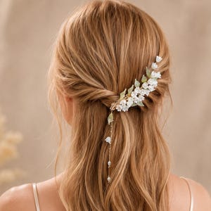 Può includere: Un accessorio per capelli con un grappolo di piccoli fiori bianchi a forma di campana e foglie verdi traslucide disposte in un design curvo. Una sottile catenina dorata pende dal pezzo, impreziosita da due piccole perline bianche.