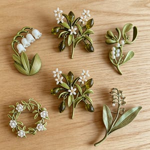 May include: A collection of six floral brooches in various designs. The brooches feature white flowers, green leaves, and accents, with a mix of styles including a wreath, sprigs, and bell-shaped flowers. The brooches are made of metal and have a vintage aesthetic.