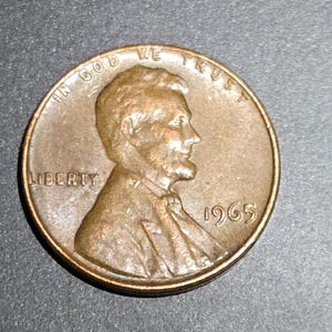 May include: A copper-colored 1965 Lincoln penny. The obverse features a profile of Abraham Lincoln, the word "LIBERTY", and the year "1965". The coin also has the inscription "IN GOD WE TRUST".