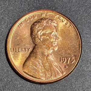 May include: A copper-colored 1975 United States penny. The obverse features a profile of Abraham Lincoln, the word "LIBERTY", and the year "1975". The coin also has the inscription "IN GOD WE TRUST".