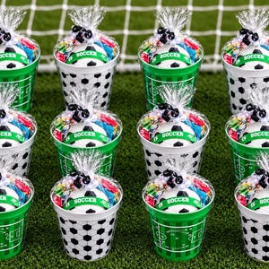 May include: Twelve plastic party favor buckets decorated with soccer ball patterns and field markings. Each bucket contains a small soccer ball with a green band labeled SOCCER, wrapped in clear cellophane with a black and white ribbon tie.